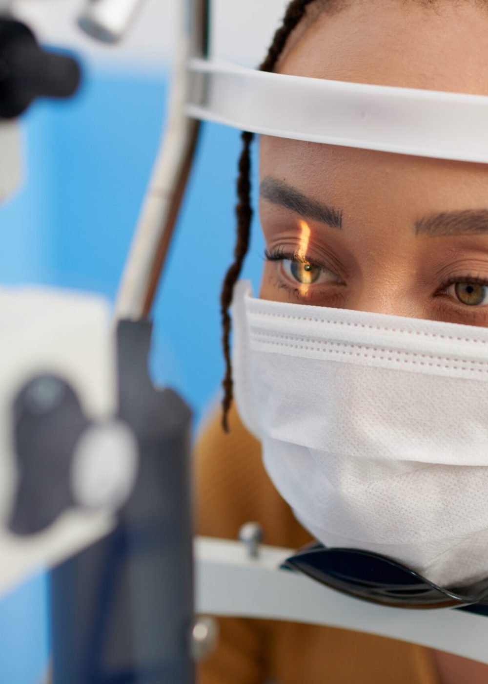 Female patient in medical mask getting eyes examined with slit lamp, microscope that allows doctor to closely examine the eye